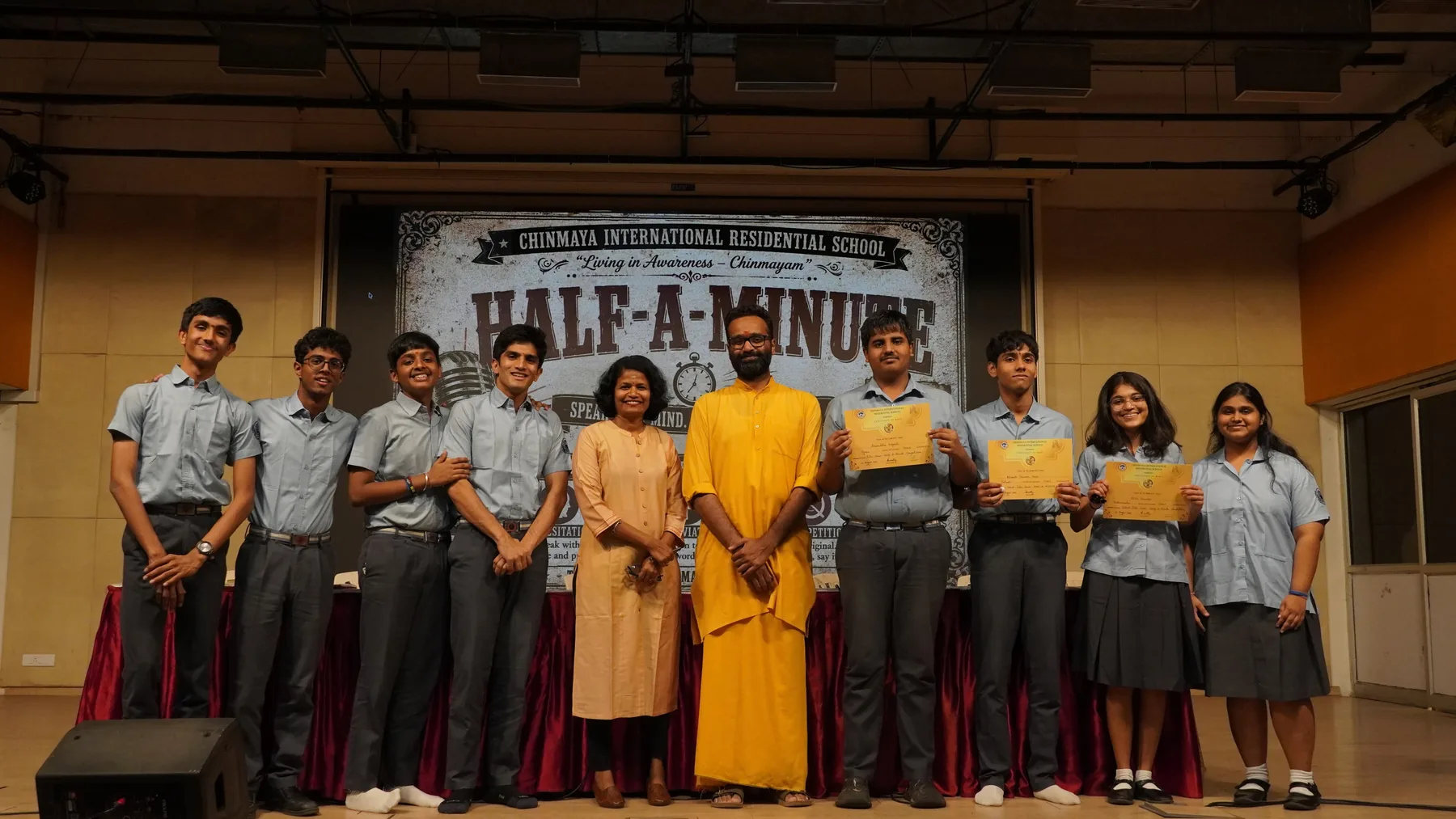 Students in school uniform and a swami in saffron standing on a stage before a Half-A-Minute banner