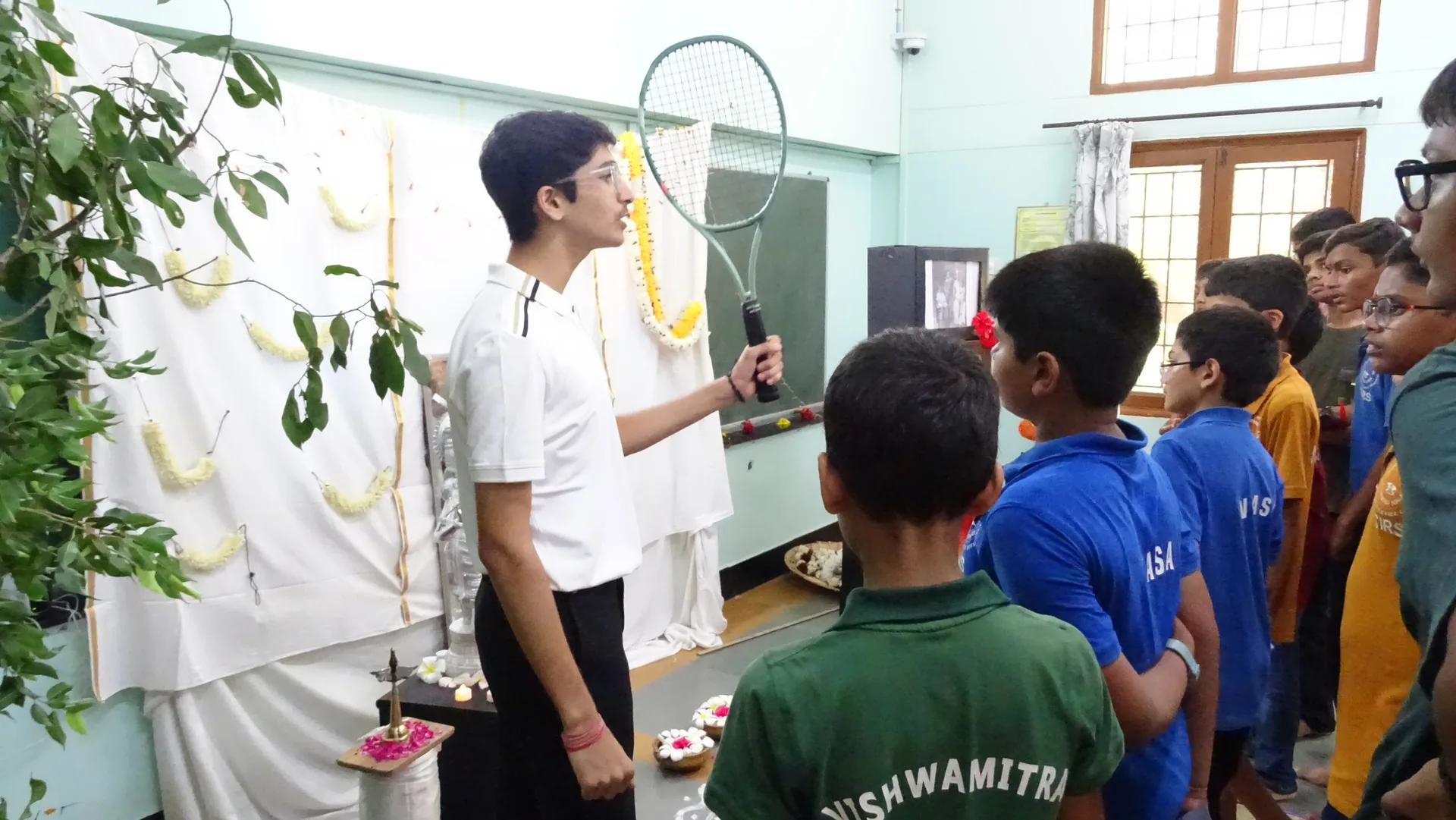 A teacher speaking into a microphone beside a decorated board as students in house shirts look on