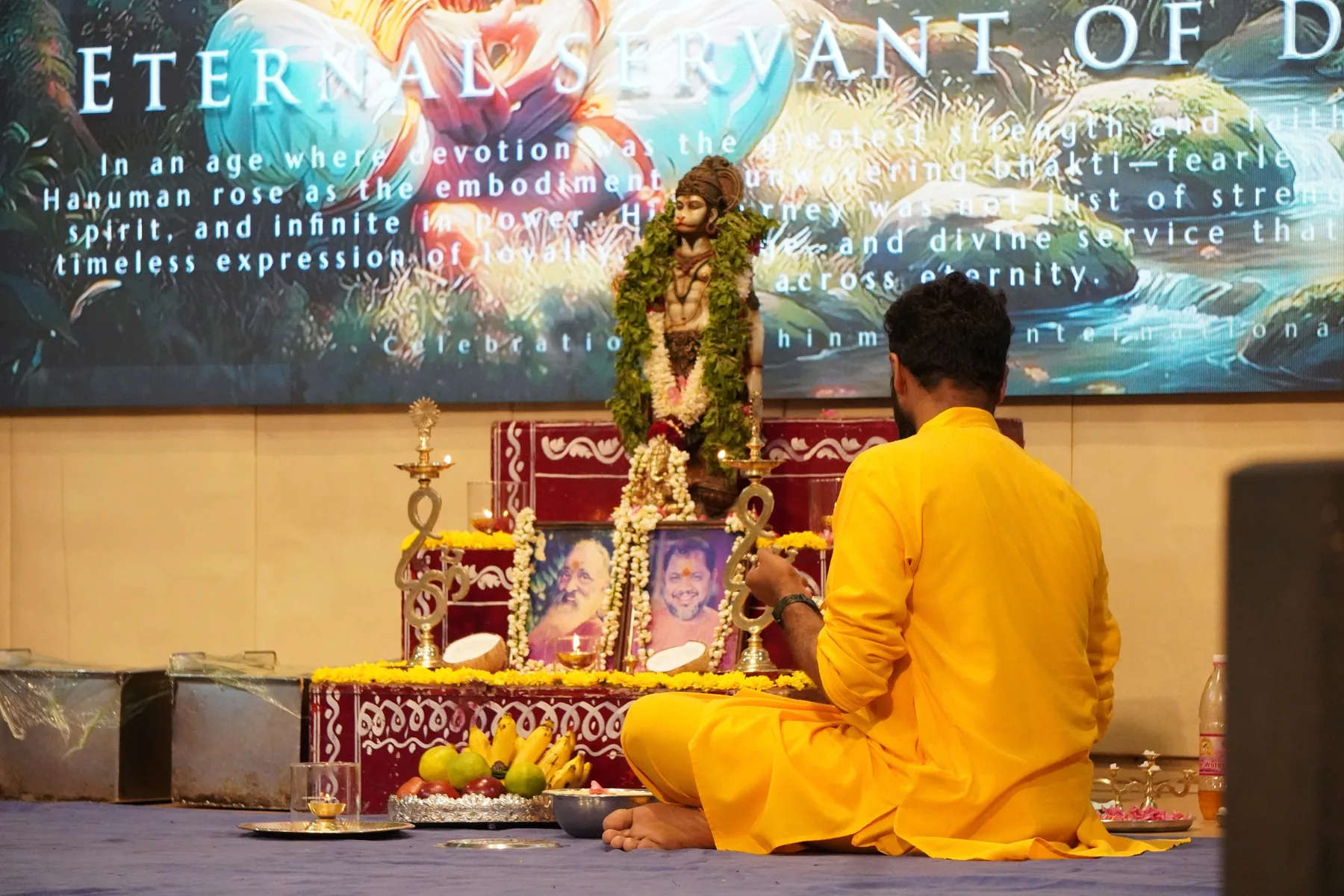 A student in a yellow kurta seated before a garlanded murti, lamps and offerings