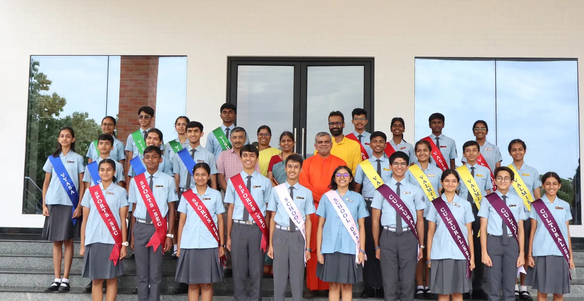 The Student Council 2026–27 standing in rows on the steps of a school building, in sashes over school uniform, with a swami in saffron among them