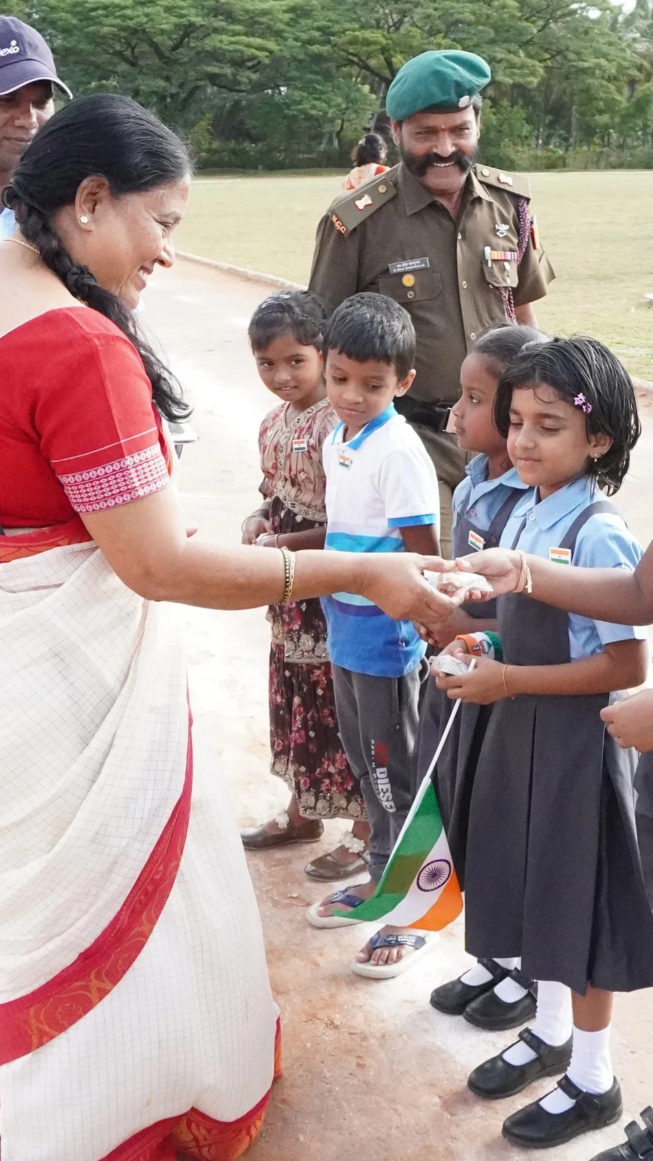 A woman in a white and red sari handing something to small children, one of them holding a flag