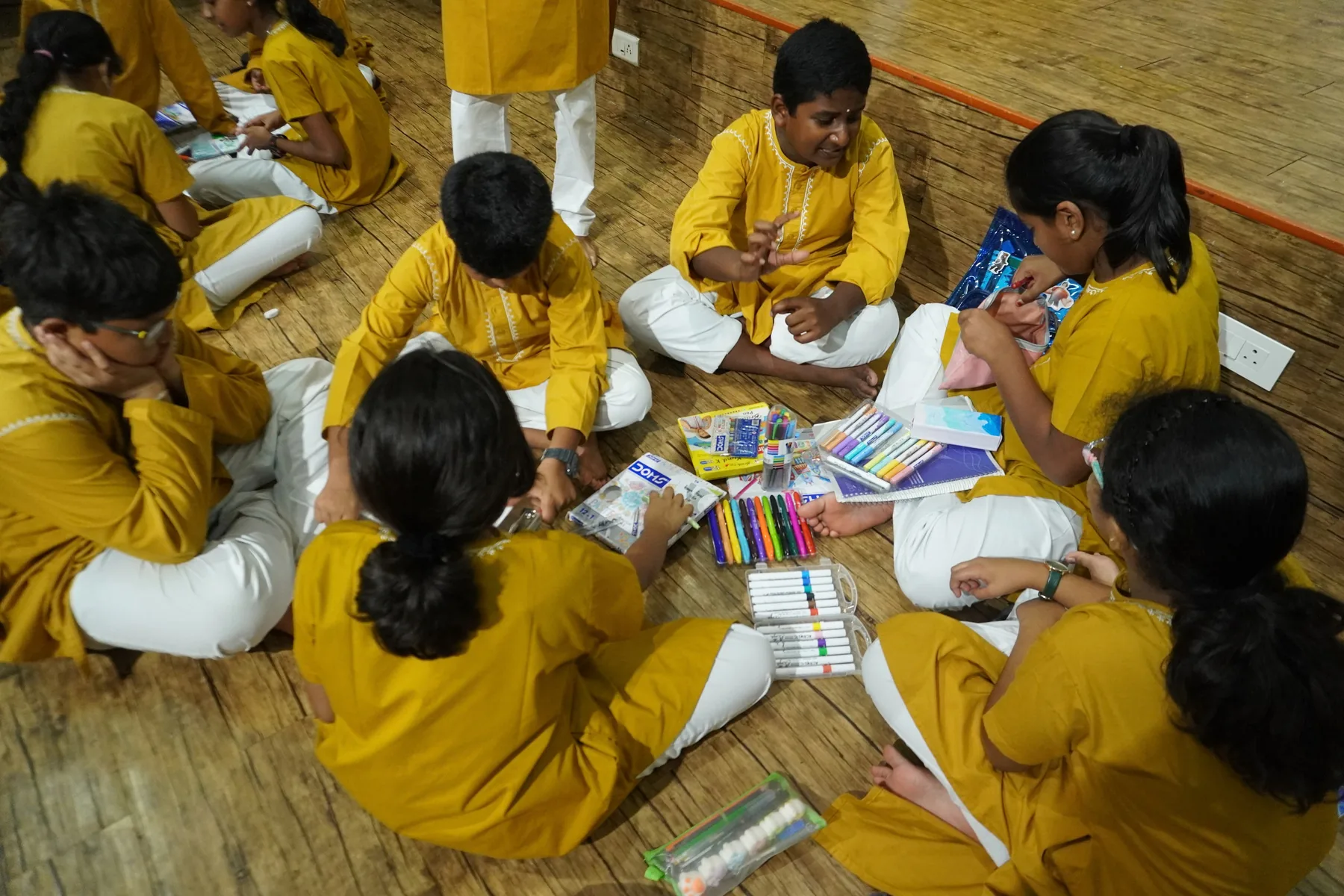 Students seated on a wooden floor with coloured pens and paper