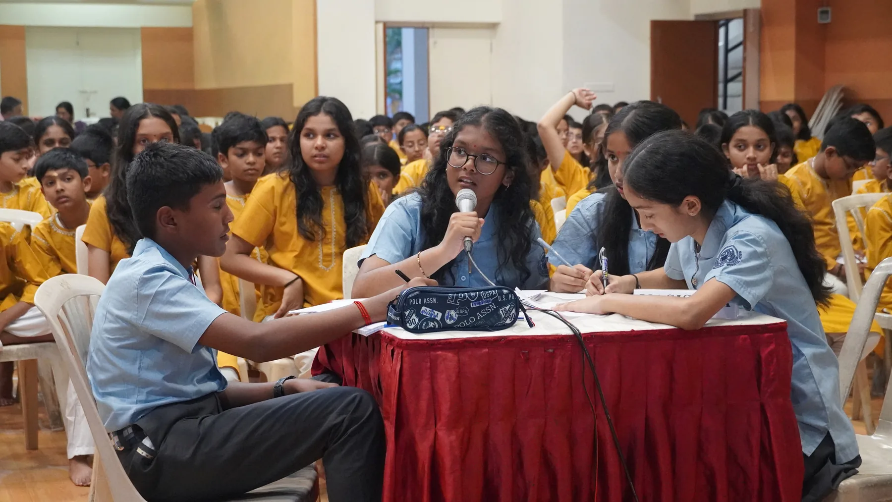Students seated at a table covered in a red cloth, answering questions before an audience