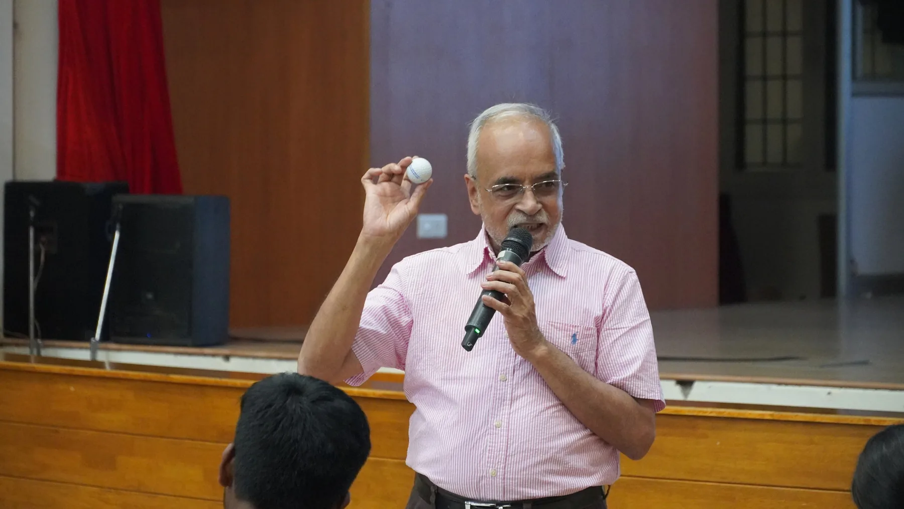 A man in a pink shirt speaking into a microphone while holding up a small white object