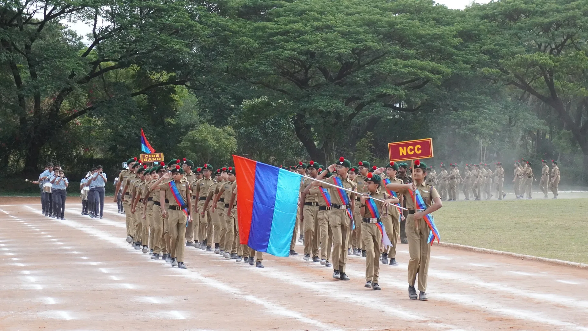 NCC cadets marching behind a blue and red flag on a parade ground