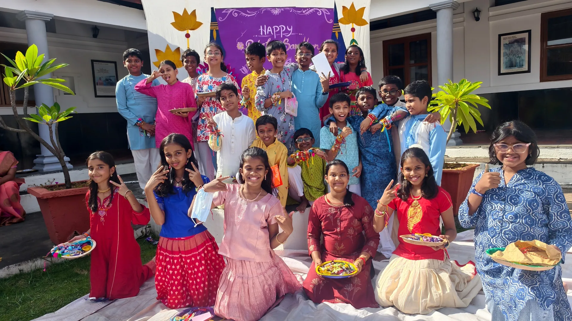 Students in festive clothes kneeling on the grass holding plates, with a group standing behind them