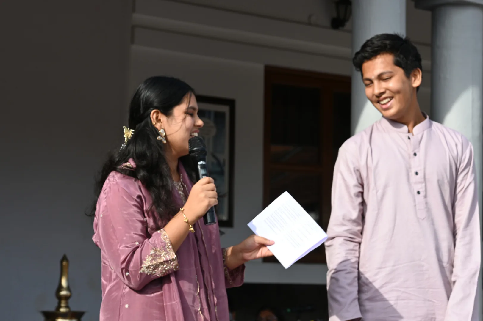 A girl speaking into a microphone beside a boy in a kurta
