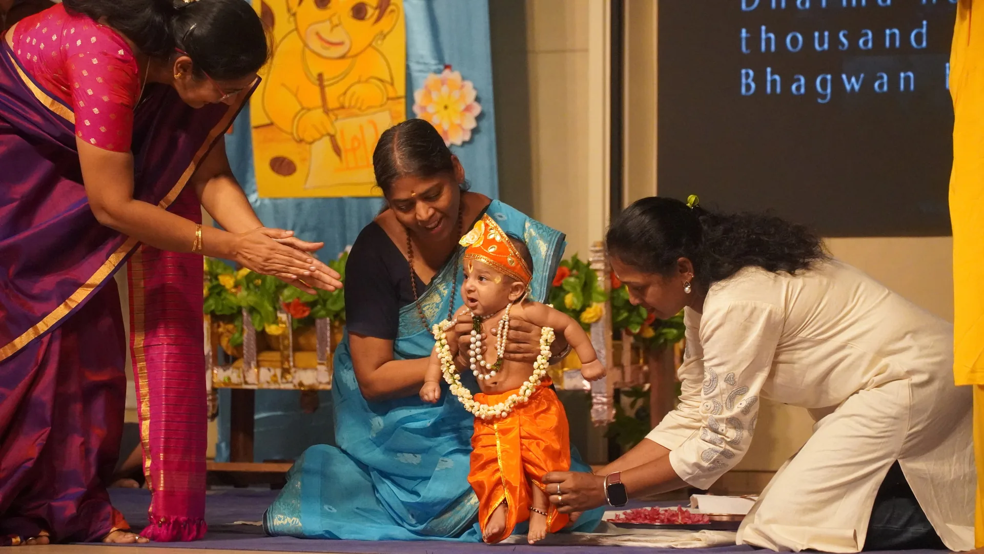 A garlanded child dressed as Bal Ram seated at a decorated altar as women make offerings