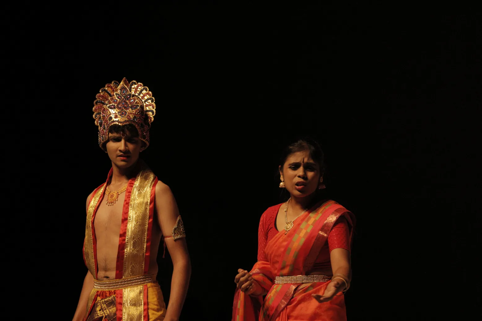 A student in a golden crown and a student in a red sari on a dark stage