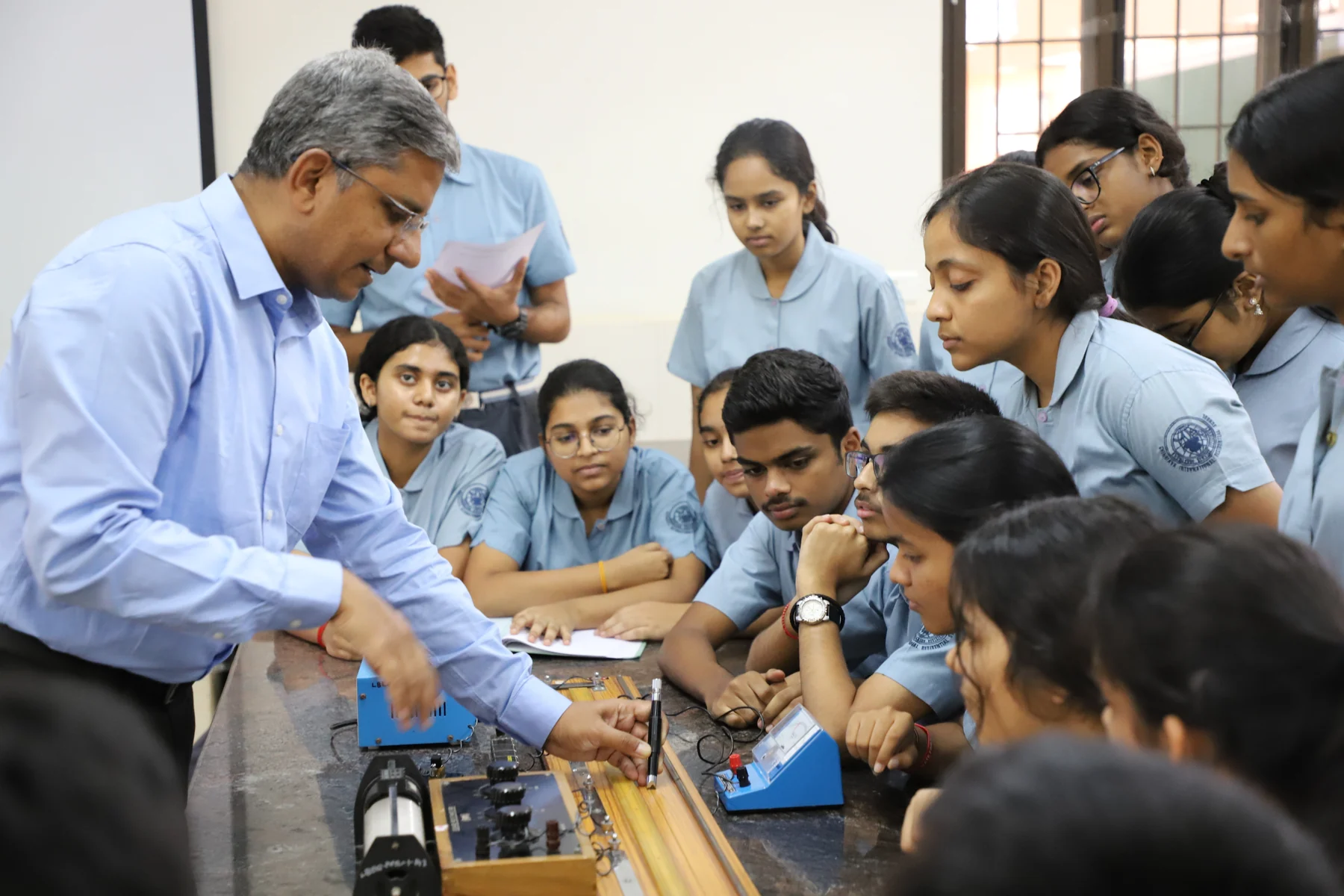 A teacher demonstrating apparatus to students gathered round a laboratory bench