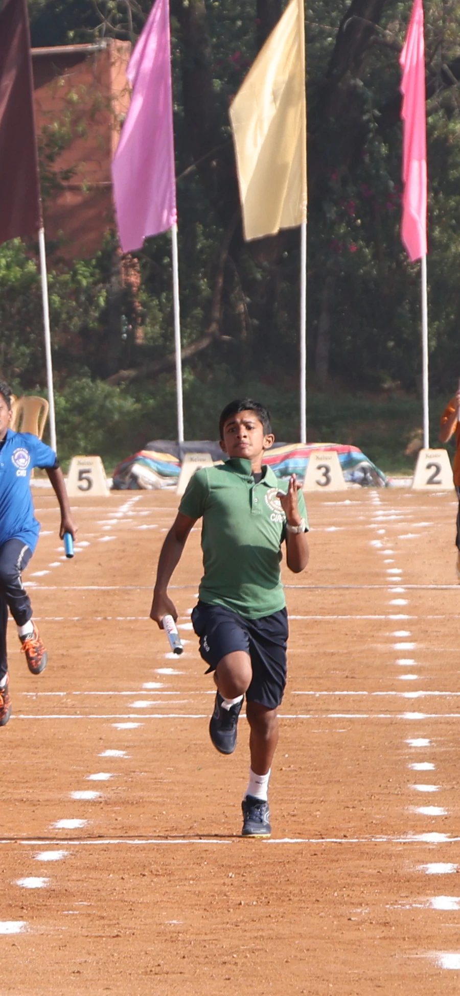 A student in a green jersey runs with a relay baton on the track, with other runners behind him.