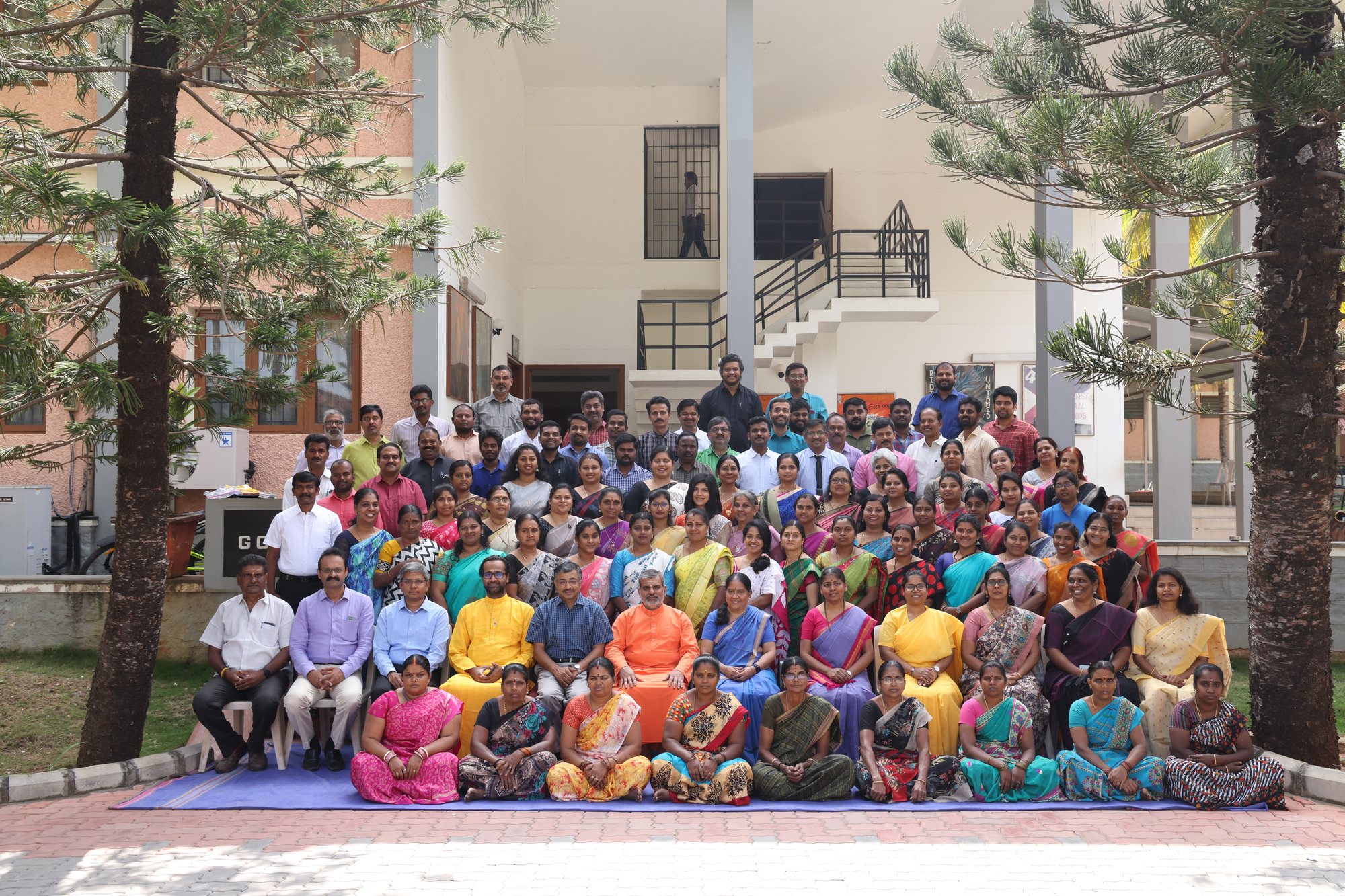 About ninety members of staff and faculty, seated and standing in rows on the steps of a campus building, between two pine trees
