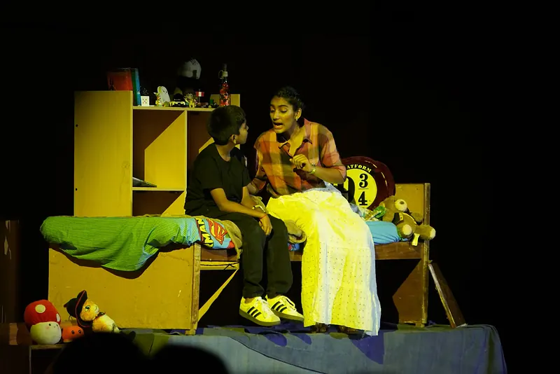 Two actors on a bed in a room set with yellow shelves and soft toys
