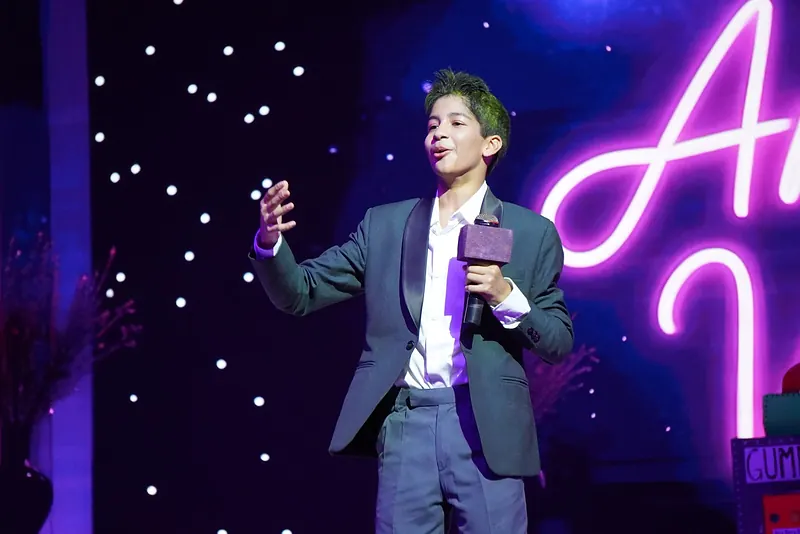 An actor in a grey suit speaking into a microphone beside a neon sign