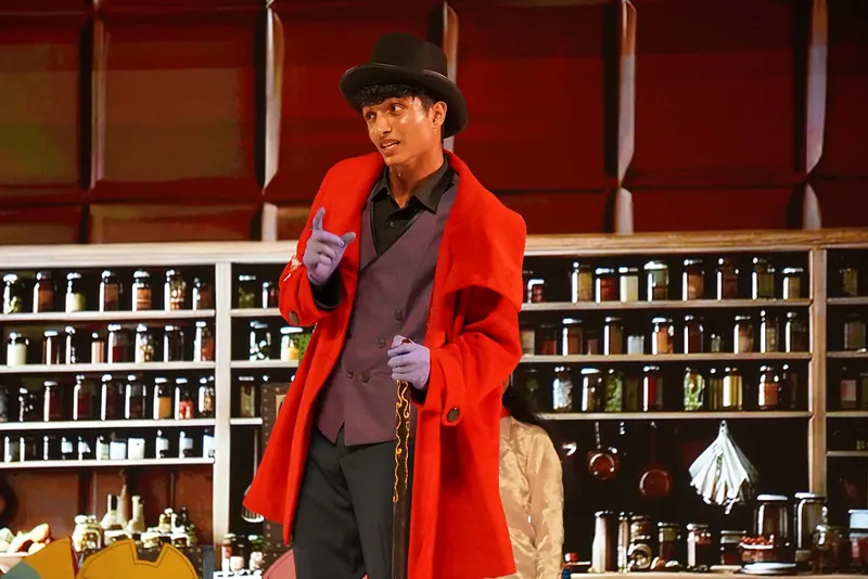 An actor in a red coat and top hat before shelves of jars