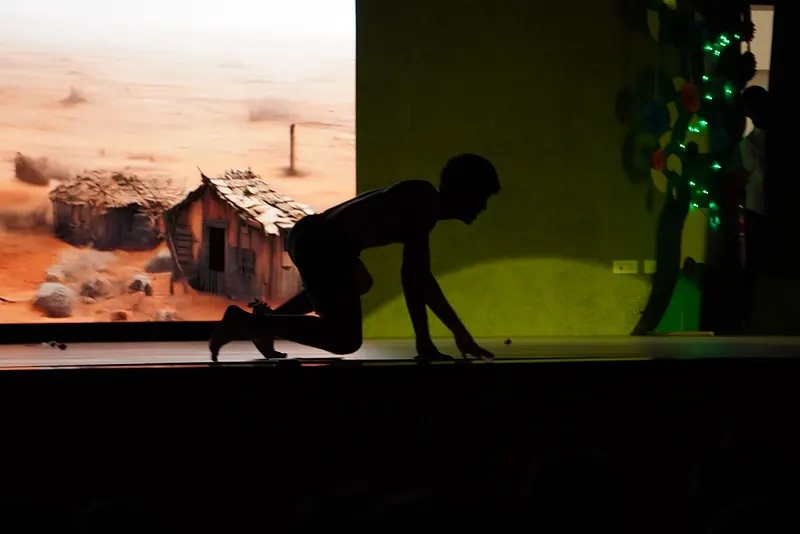 A figure crawling in silhouette before a projected desert and a ruined hut