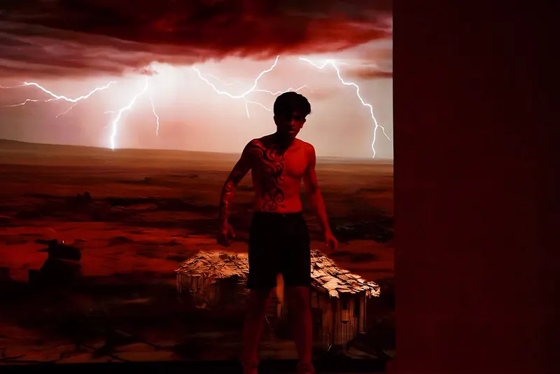 An actor in body paint standing before a projected lightning storm and a broken hut