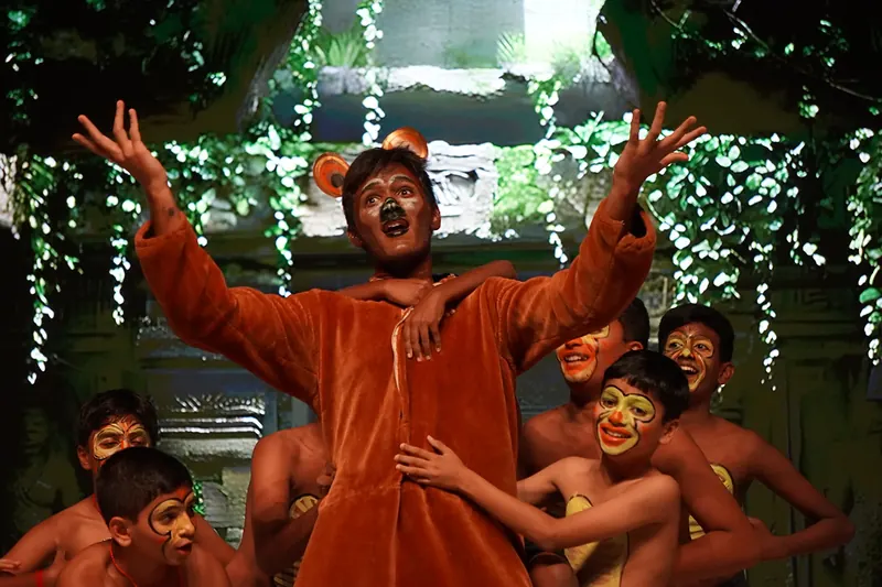 An actor in a bear costume, arms raised, among painted jungle children before a ruined temple set
