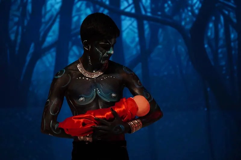 A figure in dark body paint cradling a bundle in red cloth before a blue forest