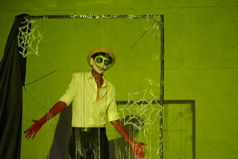 An actor in skull face paint in a green set strung with cobwebs