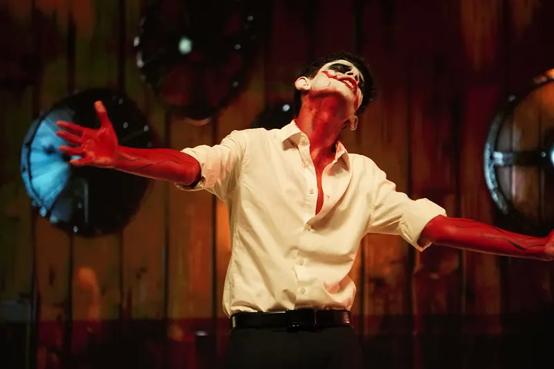 An actor in skull face paint with red-painted hands, arms spread, before a wooden wall hung with shields