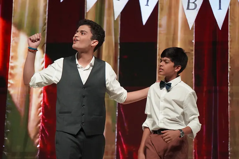 An actor in a waistcoat with a fist raised beside a younger actor in a bow tie, bunting above