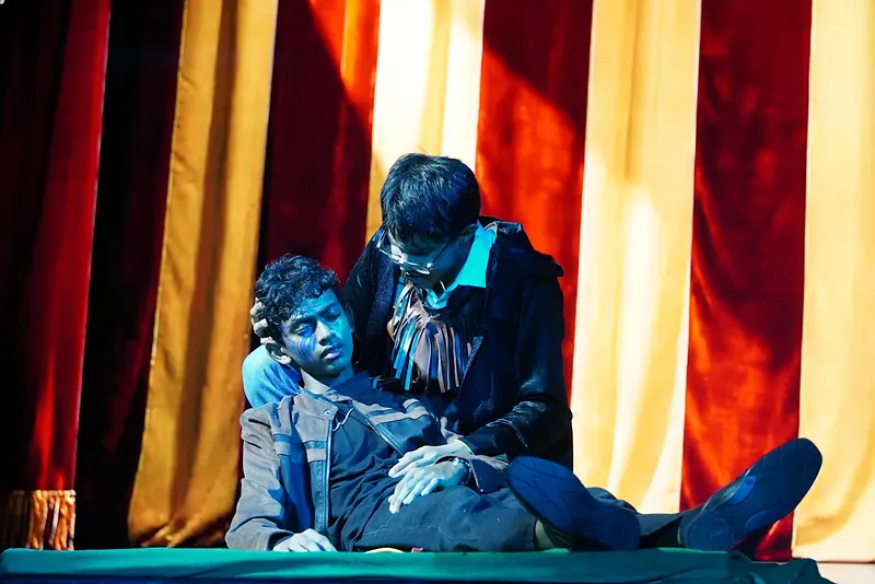 An actor kneeling over another who lies on the floor, before striped curtains