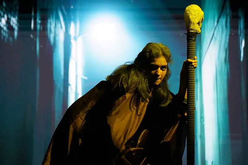 A figure in a shaggy cloak holding a skull-topped staff, lit green and gold