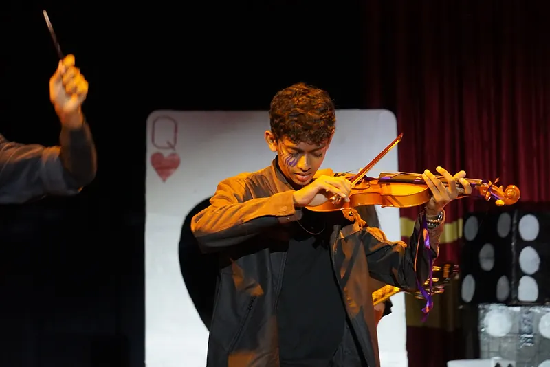 An actor playing a violin before a giant playing card
