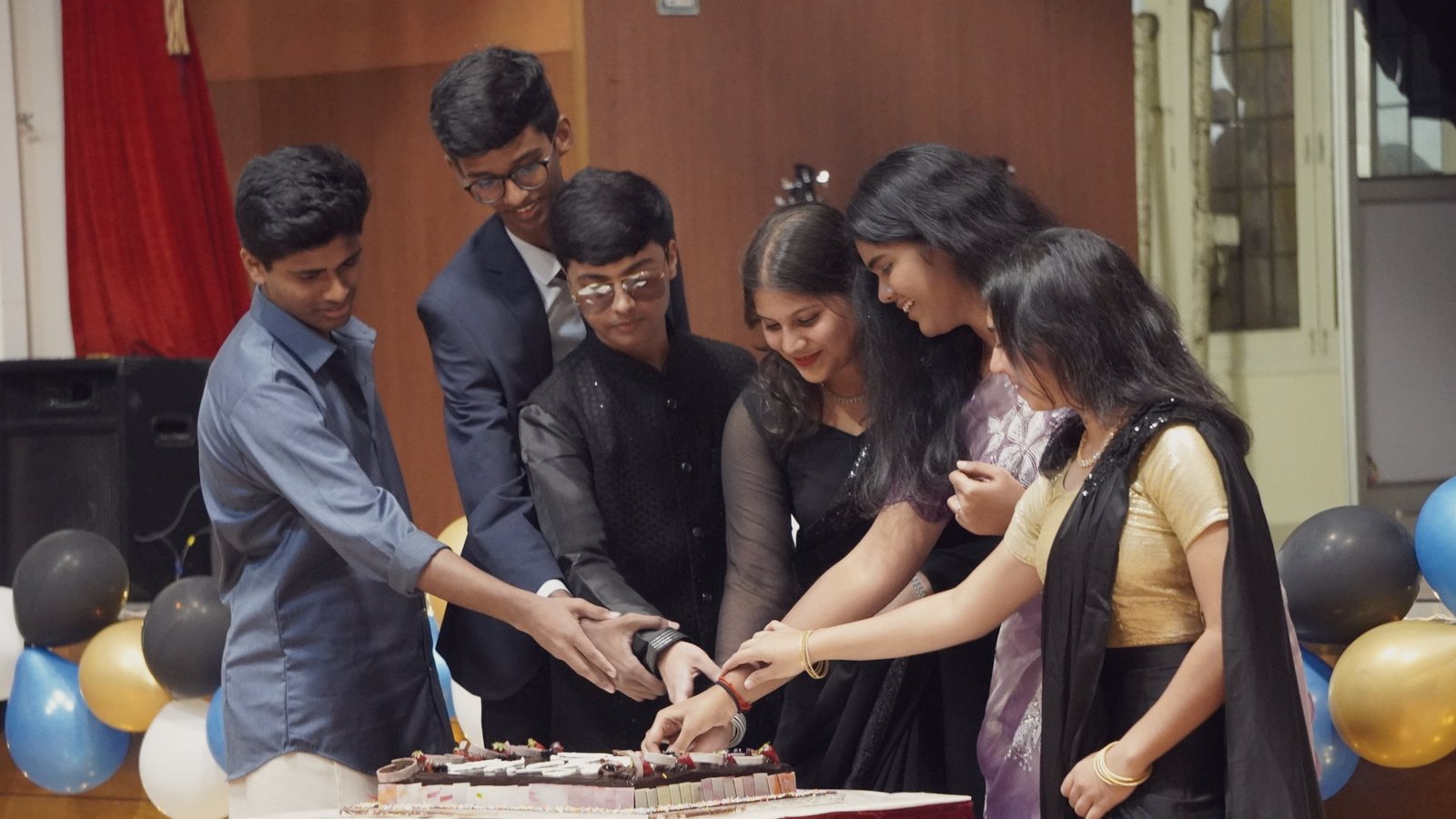 Six senior students in formal dress cutting a cake together, their hands joined on the knife