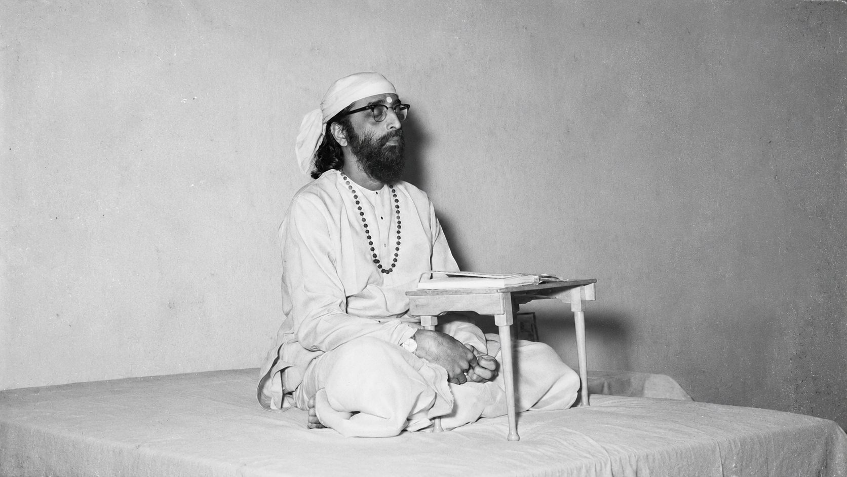Swami Chinmayananda seated in reflection, with a book resting on a low wooden table